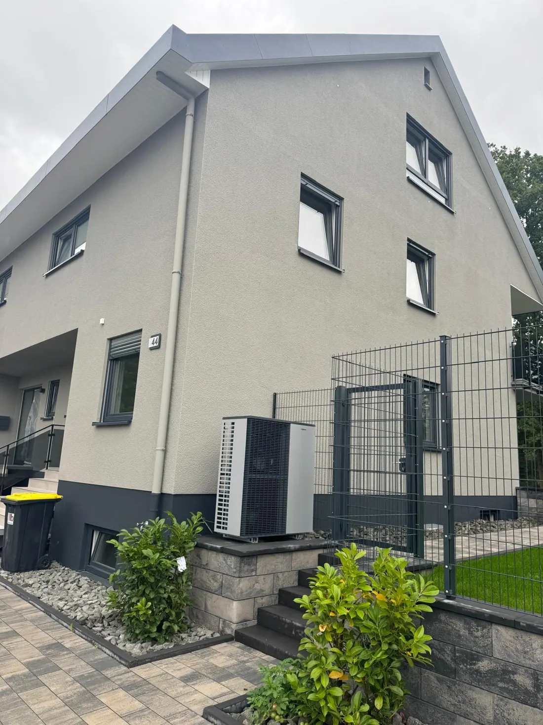 Wärmepumpe neben dem Gartentor eines Hauses mit grauer Fassade